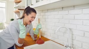 Woman using a plunger in a kitchen sink, expressing frustration over a plumbing issue, highlighting the need for plumbing services in Lazy Lake, Florida.