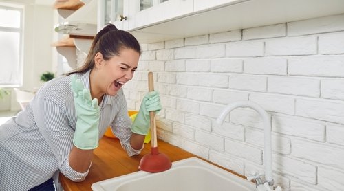 Woman using a plunger in a kitchen sink, expressing frustration over a plumbing issue, highlighting the need for plumbing services in Lazy Lake, Florida.